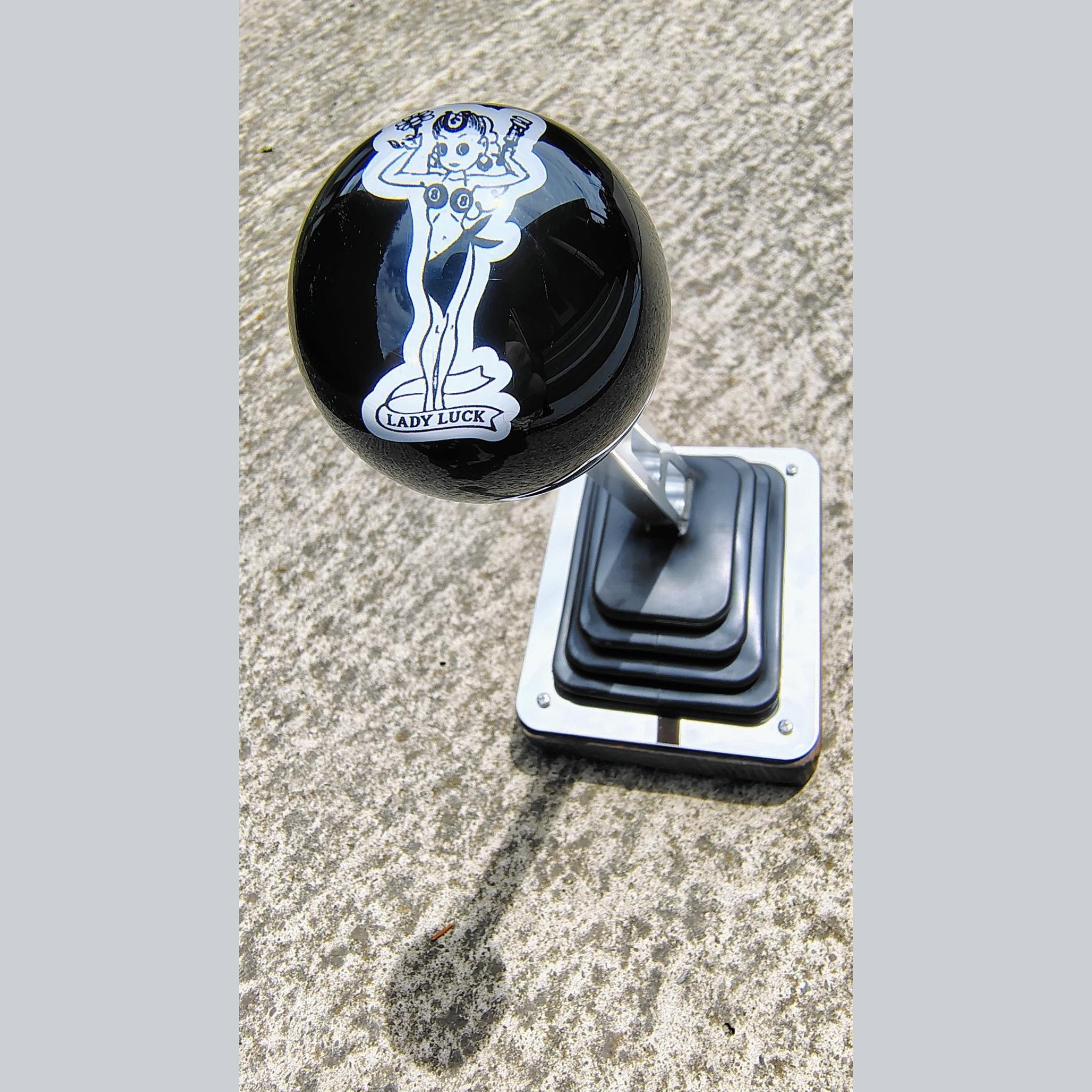 Lady Luck resin shifter ball installed on a vehicle shifter — angled overhead view showing the white-on-black vintage Lady Luck pinup artwork and banner on a deep gloss black resin ball. Chrome shift boot plate below on concrete surface. 