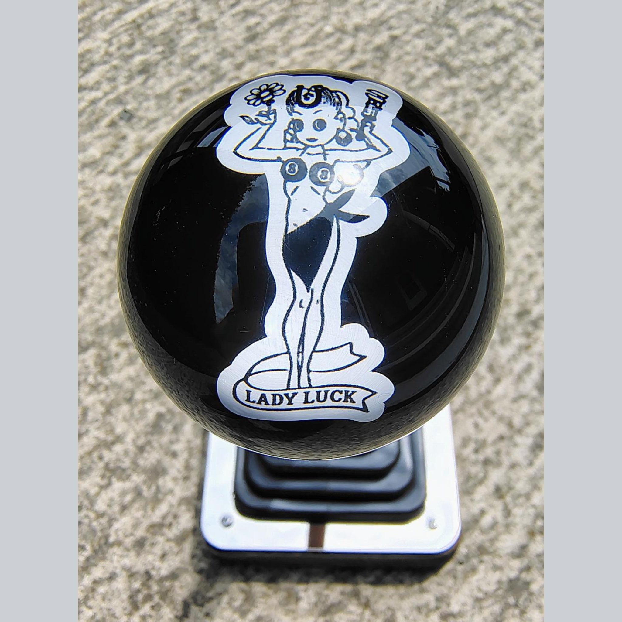 Front view of the Lady Luck resin shifter ball on a shifter — deep gloss black resin with vintage pinup girl artwork in white, holding a flower and wrench, with 'Lady Luck' banner below. Crystal-clear resin dome finish. 