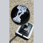 Side-angle view of the Lady Luck resin shifter ball mounted on a chrome shift boot plate. Deep gloss black ball with white Lady Luck vintage pinup artwork visible on the side. Shot from slightly below on outdoor concrete surface.