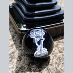 Lady Luck handmade resin shifter ball resting on concrete with shift boot plate in background — gloss black resin body showing the white vintage Lady Luck pinup artwork and banner. Uninstalled display view.