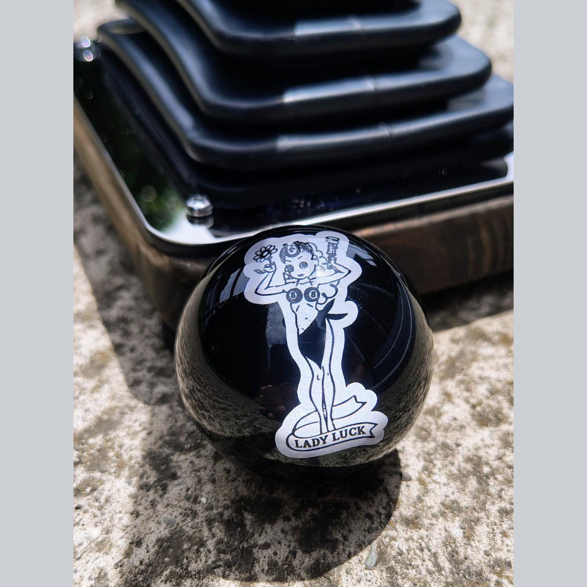 Lady Luck handmade resin shifter ball resting on concrete with shift boot plate in background — gloss black resin body showing the white vintage Lady Luck pinup artwork and banner. Uninstalled display view.