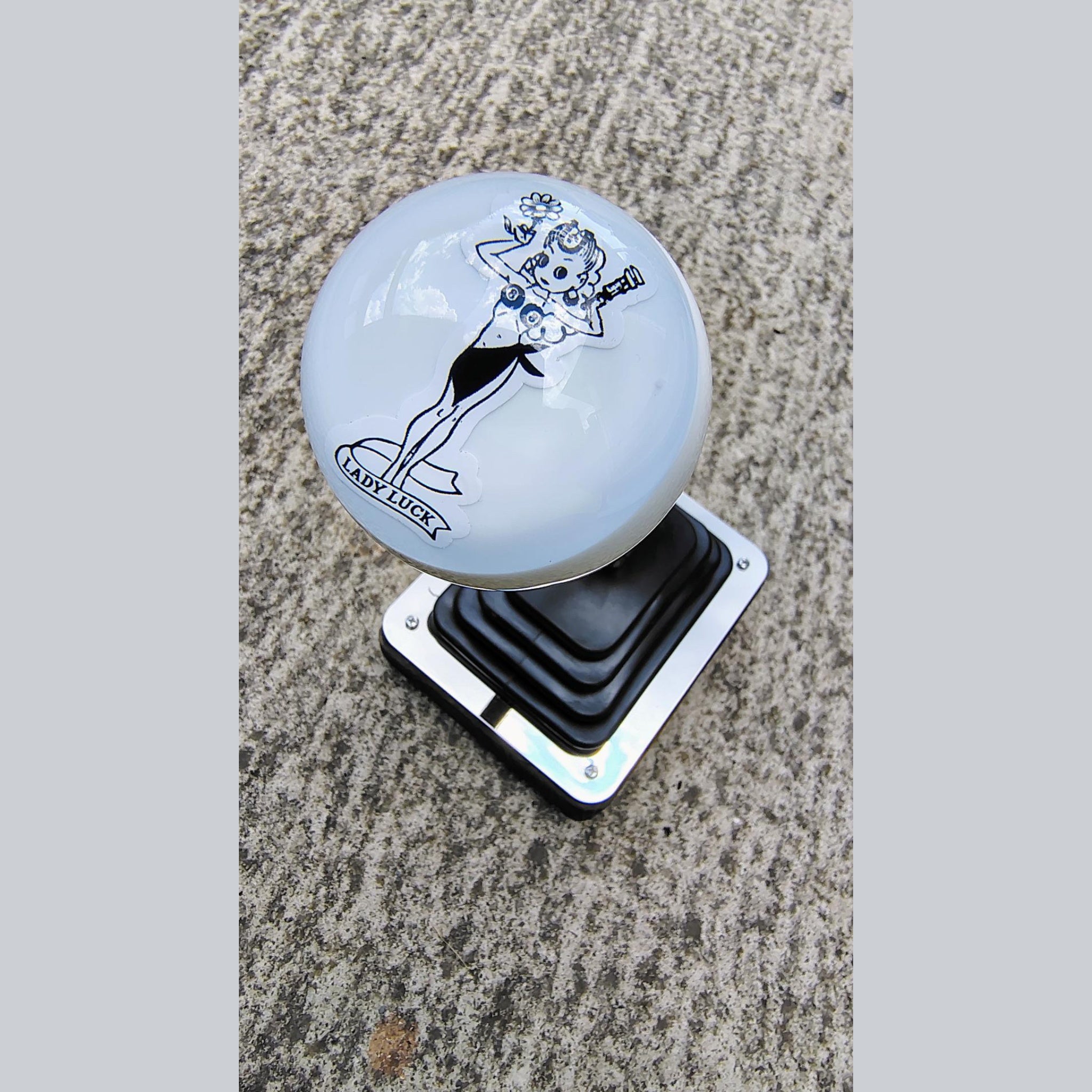 Lady Luck resin shifter ball installed on a vehicle shifter — angled overhead view showing the black-on-white vintage Lady Luck pinup artwork and banner on a gloss white resin ball. Chrome shift boot plate below on concrete surface. 