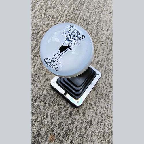 Lady Luck resin shifter ball installed on a vehicle shifter — angled overhead view showing the black-on-white vintage Lady Luck pinup artwork and banner on a gloss white resin ball. Chrome shift boot plate below on concrete surface. 