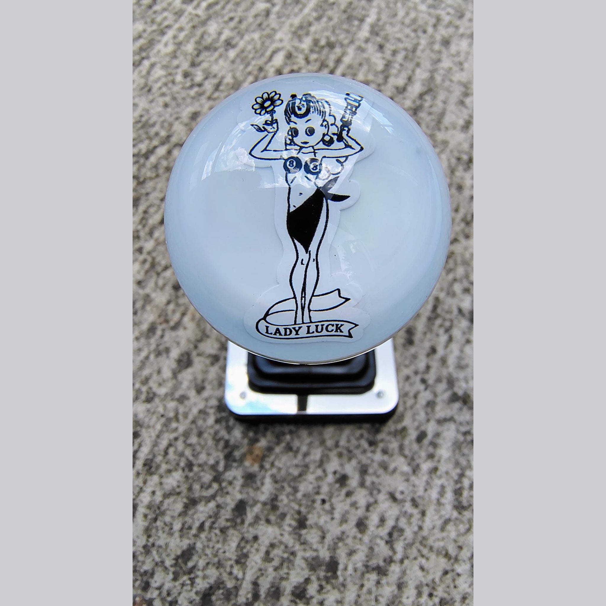 Front view of the Lady Luck resin shifter ball on a shifter — gloss white resin with vintage pinup girl artwork in black, holding a flower and wrench, with 'Lady Luck' banner below. Crystal-clear resin dome finish.