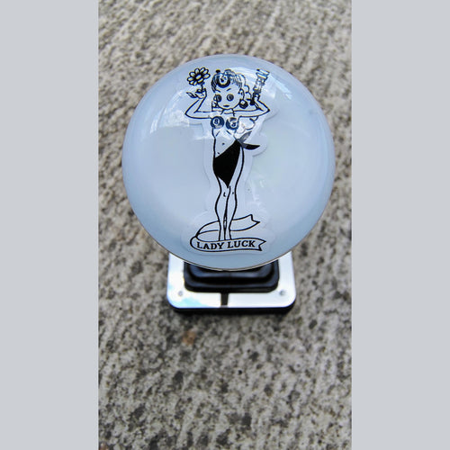 Front view of the Lady Luck resin shifter ball on a shifter — gloss white resin with vintage pinup girl artwork in black, holding a flower and wrench, with 'Lady Luck' banner below. Crystal-clear resin dome finish.