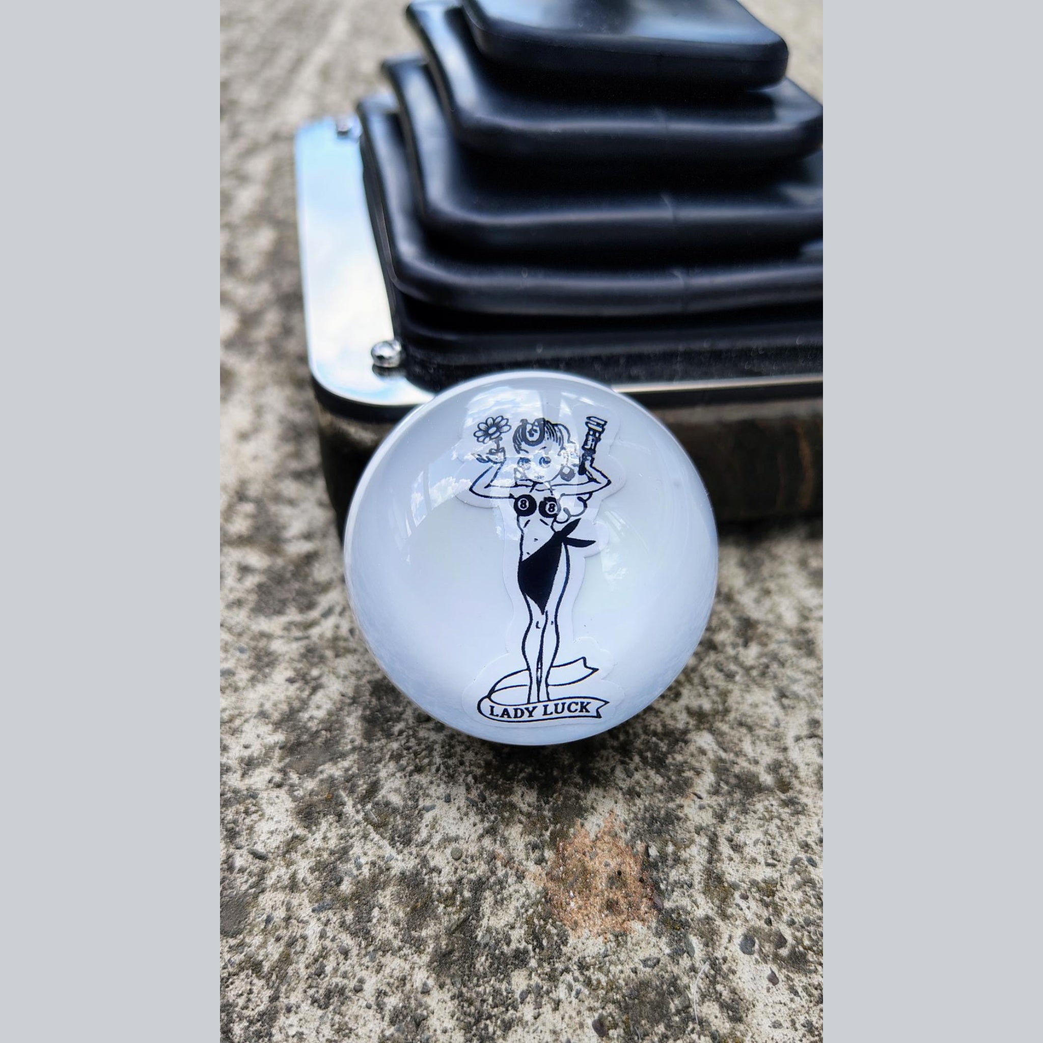 Lady Luck handmade resin shifter ball resting on concrete with shift boot plate in background — gloss white resin body showing the black vintage Lady Luck pinup artwork and banner. Uninstalled display view.