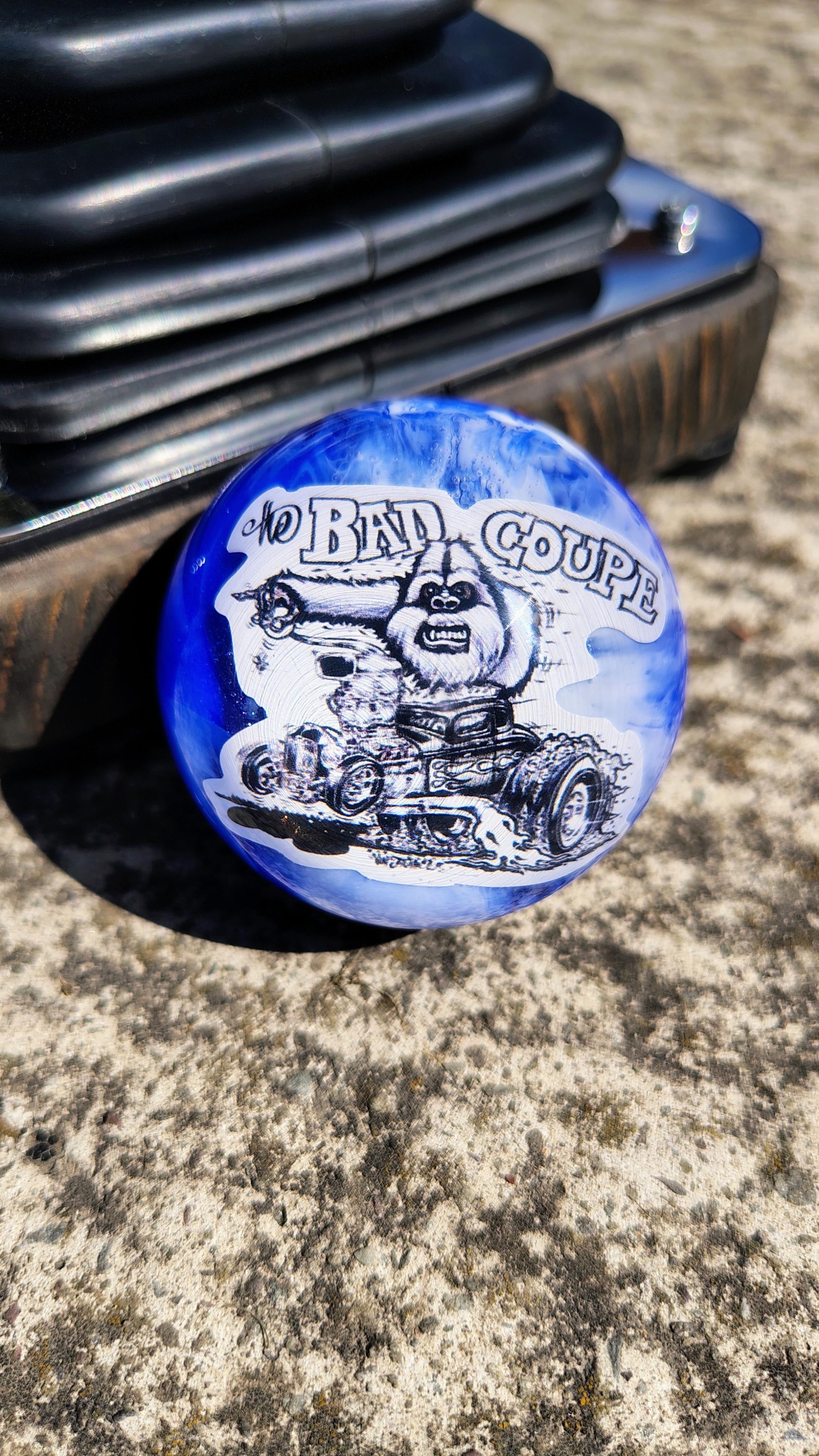 The Bad Coupe Resin Shifter Ball in swirled blue and white resin, showing Von Franco's hand-drawn Yeti driving a blown chopped hot rod coupe beneath The Bad Coupe lettering, mounted beside a shifter boot