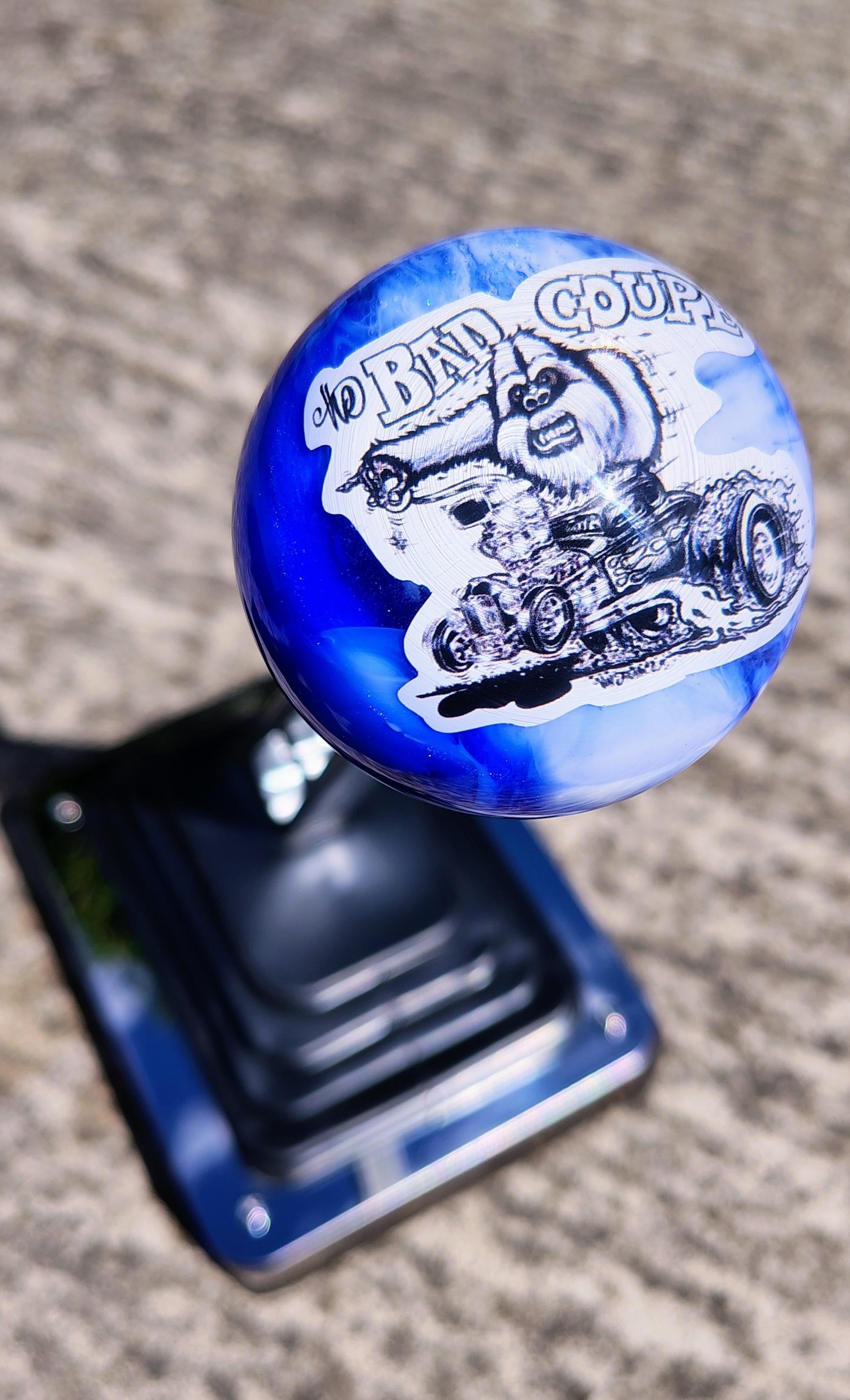 Top-down view of The Bad Coupe Resin Shifter Ball installed on a hot rod shifter, showing the full Yeti hot rod graphic and the deep cobalt blue and white marbled resin