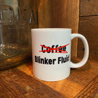 A white ceramic coffee mug with the text 'Coffee' crossed out in red and 'Blinker Fluid' printed below in black, sitting on a wooden surface next to a glass jar.