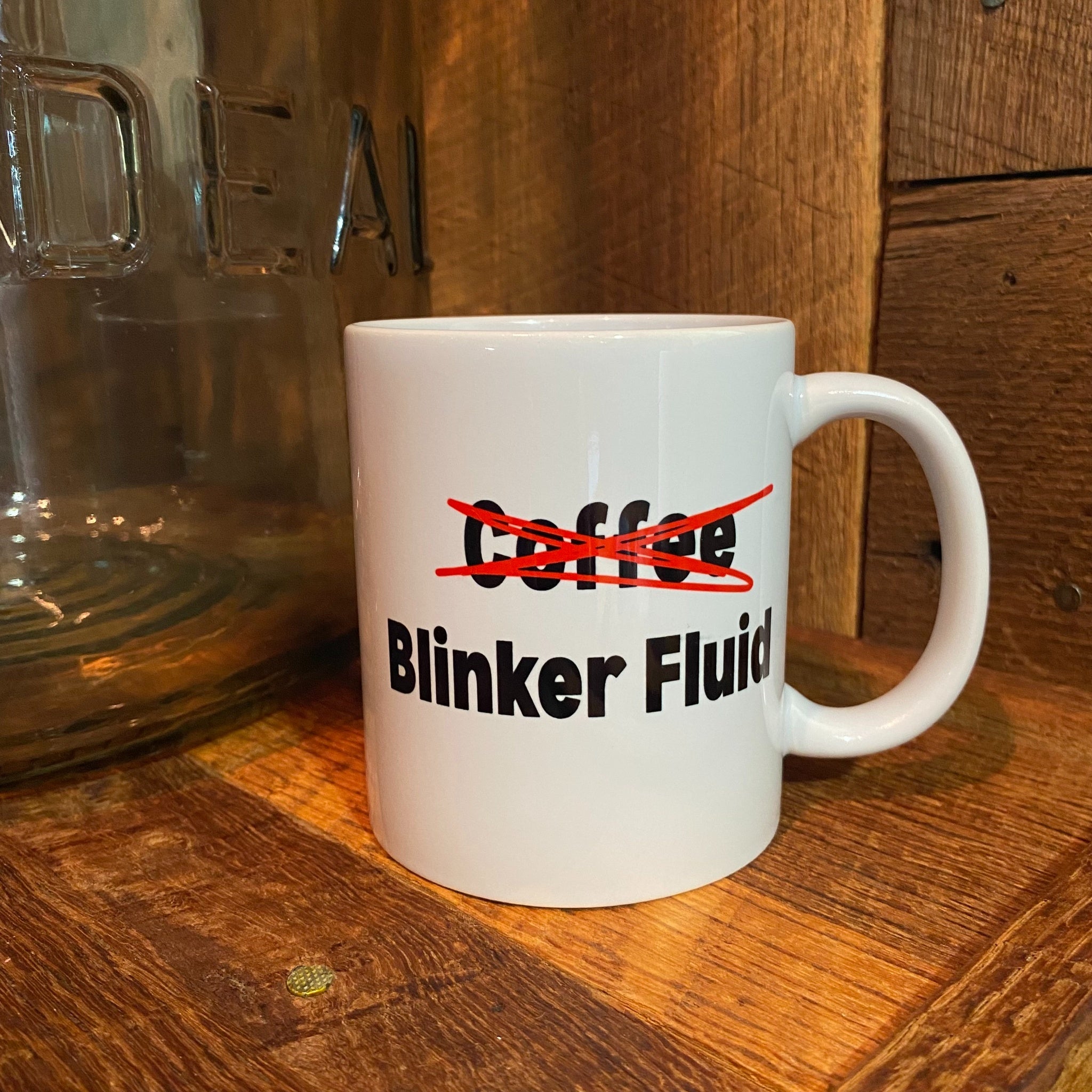 A white ceramic coffee mug with the text 'Coffee' crossed out in red and 'Blinker Fluid' printed below in black, sitting on a wooden surface next to a glass jar.