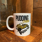 A white ceramic mug with a graphic print of a vintage car and the text 'PUDDING CREEK ROD SHOP', placed on a rustic wooden surface.