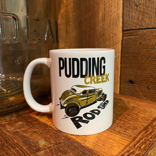 A white ceramic mug with a graphic print of a vintage car and the text 'PUDDING CREEK ROD SHOP', placed on a rustic wooden surface.