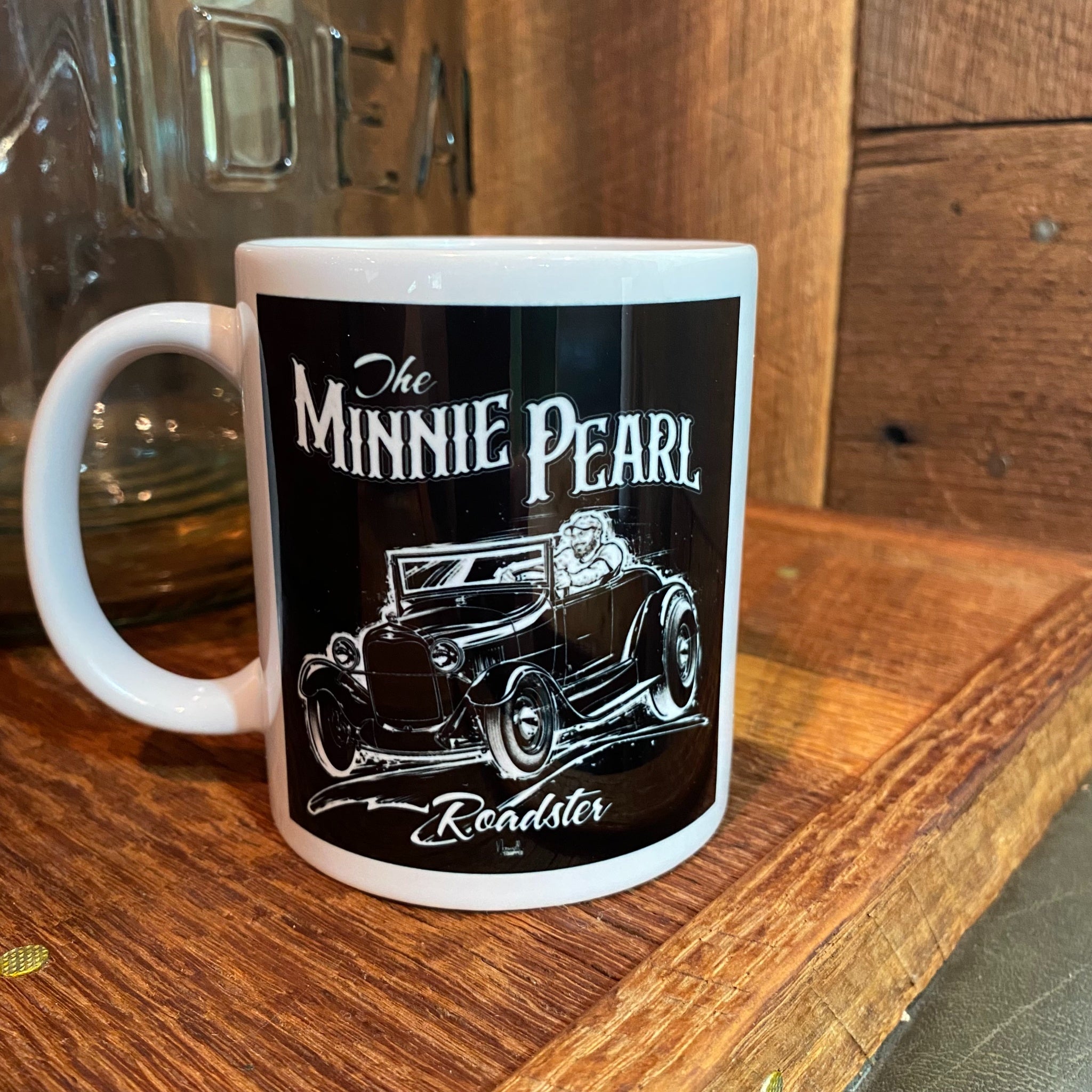 The Minnie Pearl Mug front — white ceramic coffee mug with a black wrap graphic featuring 'The Minnie Pearl' lettering above a white illustration of a 1928 Ford Roadster hot rod with driver, and 'Roadster' in script below. Displayed on a wood surface with a vintage glass jar in the background.