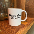 The Minnie Pearl Mug back — white ceramic coffee mug showing the Pudding Creek Rod Shop script logo in black lettering. Wood surface and vintage glass jar visible in background.