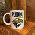 A white ceramic mug with a 'Pudding Creek Rod Shop' graphic featuring a vintage gold car, set on a rustic wooden surface next to a glass jar.