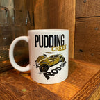A white ceramic mug with a 'Pudding Creek Rod Shop' graphic featuring a vintage gold car, set on a rustic wooden surface next to a glass jar.