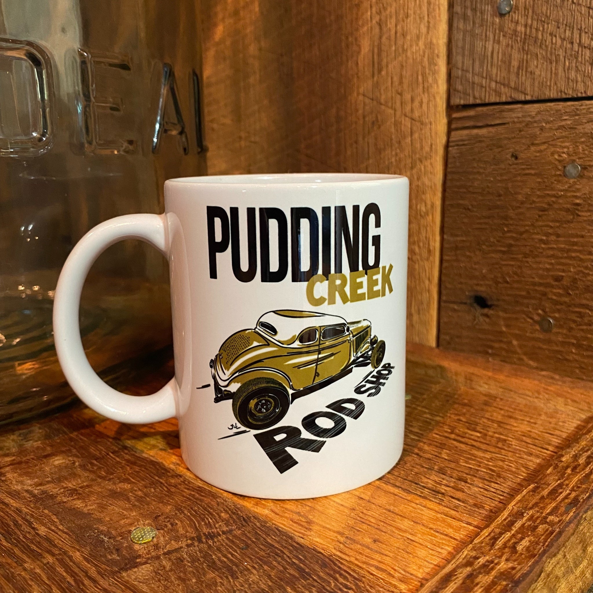 A white ceramic mug with a 'Pudding Creek Rod Shop' graphic featuring a vintage gold car, set on a rustic wooden surface next to a glass jar.