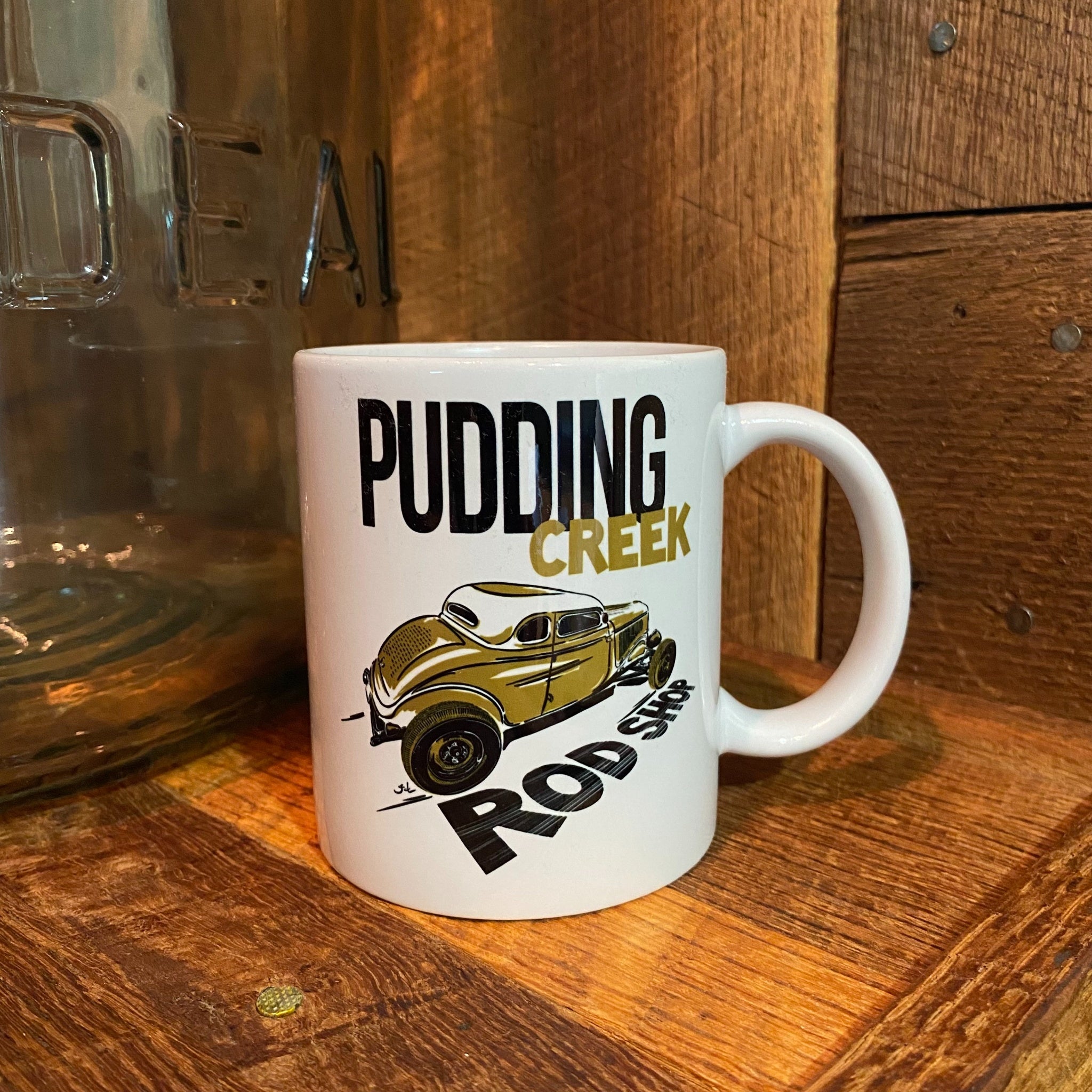 A white ceramic mug with a 'Pudding Creek Rod Shop' graphic featuring a vintage gold car, set on a rustic wooden surface next to a glass jar.