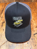 Charcoal Richardson 112 trucker hat viewed from above on a rustic wood surface, showing the full black front panel with the Pudding Creek Oregon leather logo patch featuring white block lettering, yellow "Creek" text, and a gold vintage vehicle illustration.