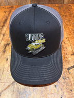 Charcoal Richardson 112 trucker hat viewed from above on a rustic wood surface, showing the full black front panel with the Pudding Creek Oregon leather logo patch featuring white block lettering, yellow "Creek" text, and a gold vintage vehicle illustration.