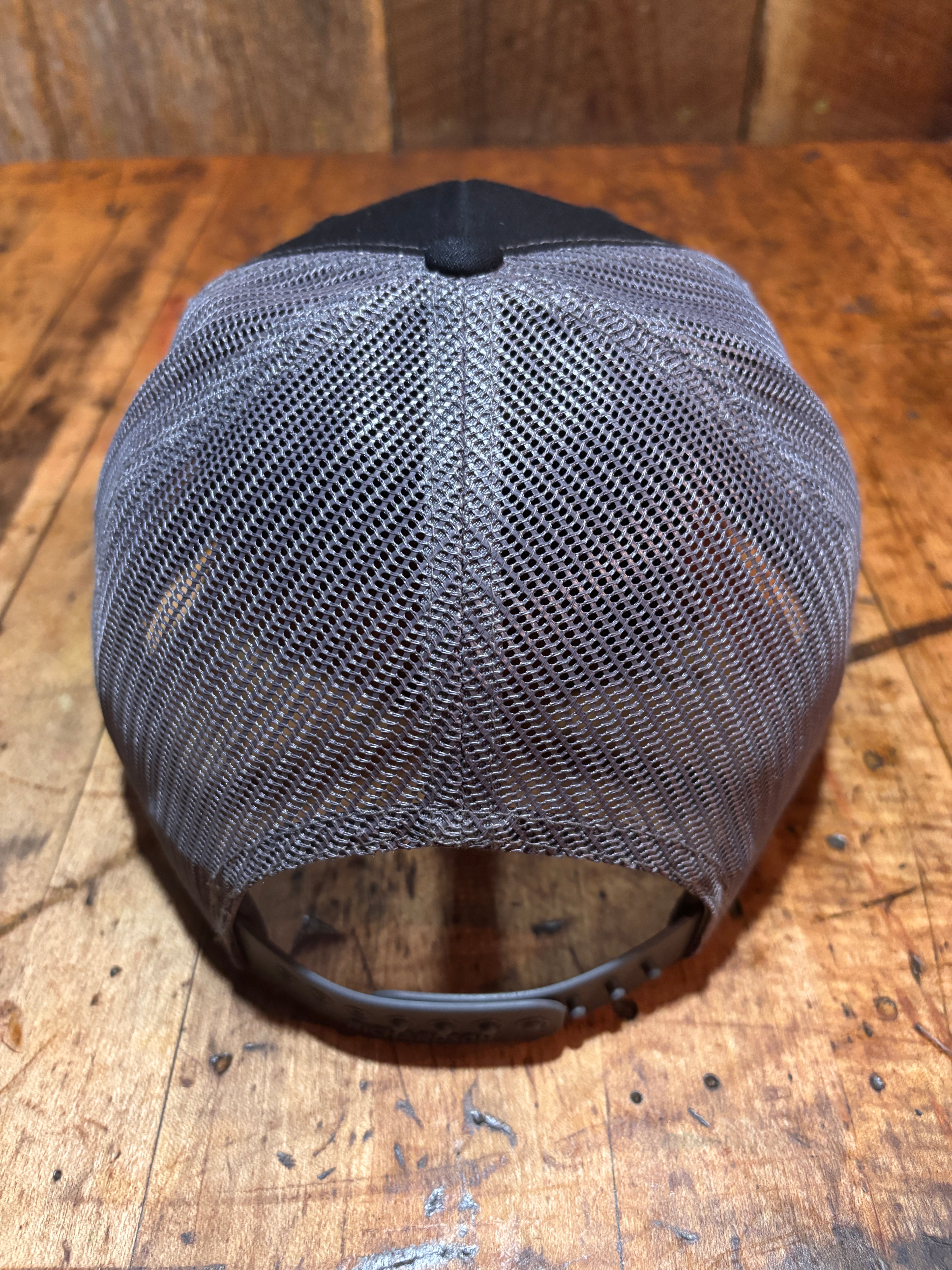 Top-down view of the Pudding Creek charcoal trucker hat showing the grey polyester mesh back panels and circular snapback closure ring, photographed on a rustic wooden surface.