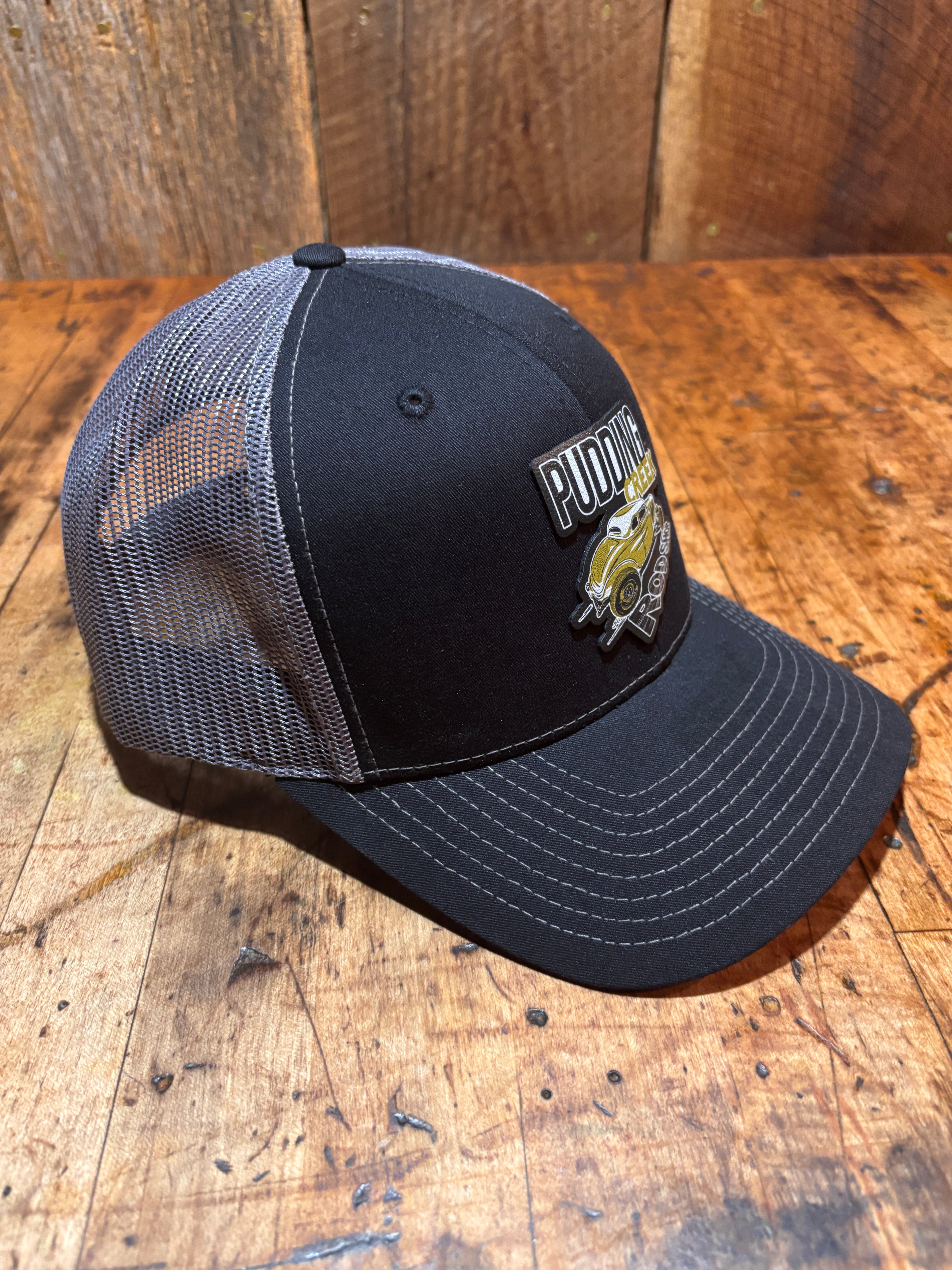Angled side view of the Pudding Creek charcoal trucker hat resting against a wooden surface, showing the black cotton-polyester front panel, grey mesh back, precurved bill with white contrast stitching, and the Pudding Creek Oregon leather patch on the crown.
