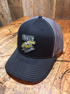 Side profile of the Pudding Creek charcoal trucker hat on a rustic wood surface, showing the black front panel, grey mesh back, and the full Pudding Creek Oregon leather patch with white "Pudding" text, yellow "Creek" lettering, and a gold illustrated vintage vehicle.