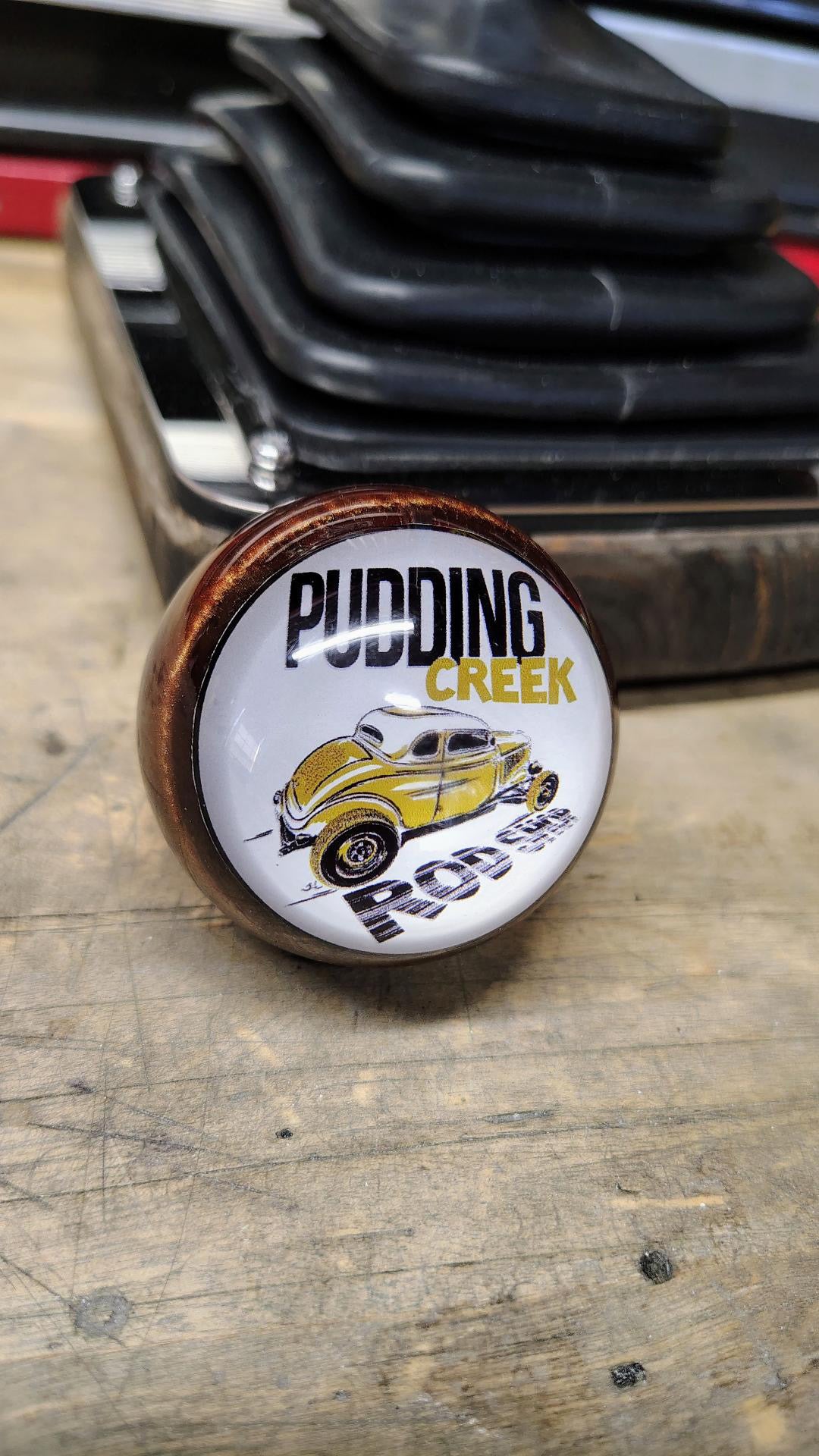 Pudding Creek Rod Shop resin shifter ball resting on a workbench, top face visible. White circular resin face displays the shop logo with 'Pudding Creek' in black and gold lettering and an illustration of a yellow vintage hot rod. Dark bronze metallic resin body.