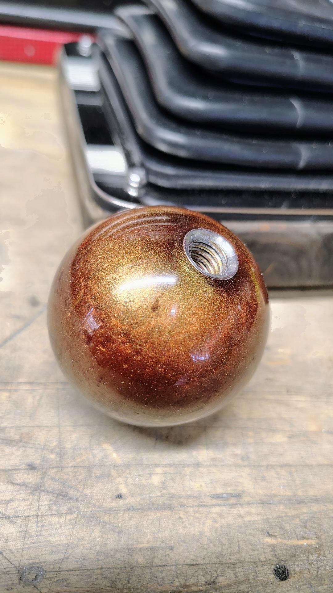 Underside of the Pudding Creek Rod Shop resin shifter ball showing the stainless steel threaded insert used for mounting on a shift lever. The ball features a swirled metallic bronze and gold resin finish with gold flake sparkle throughout.