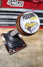 Pudding Creek Rod Shop resin shifter ball installed on a vehicle shift lever in a garage workshop. Top-down view shows the shop logo with 'Pudding Creek Rod Shop' in black and gold lettering and a yellow vintage hot rod illustration on a white resin face. Bronze metallic resin body with chrome shift boot and base plate below. Weld Racing toolbox visible in background.