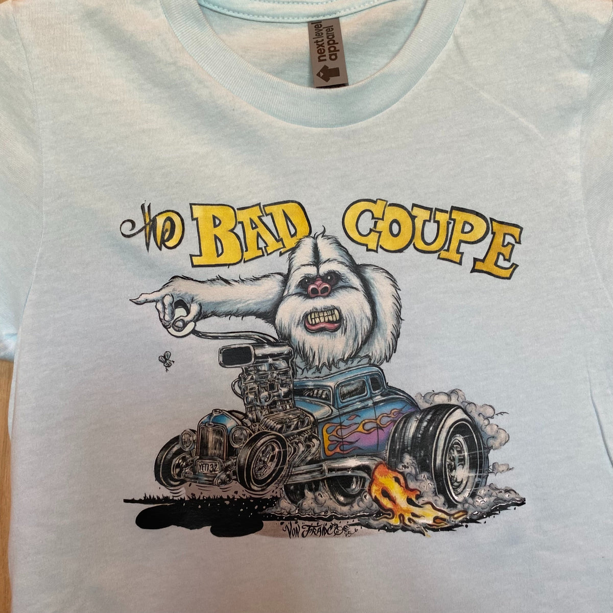 Close-up detail of The Bad Coupe Yeti graphic on the Ice Blue tee, showing the Yeti's white fur, the blown coupe engine, and the Von Franco signature, with the Next Level apparel neck tag visible