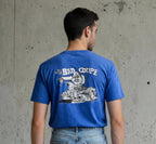 Man wearing a blue t-shirt with a graphic design on the back against a concrete wall.