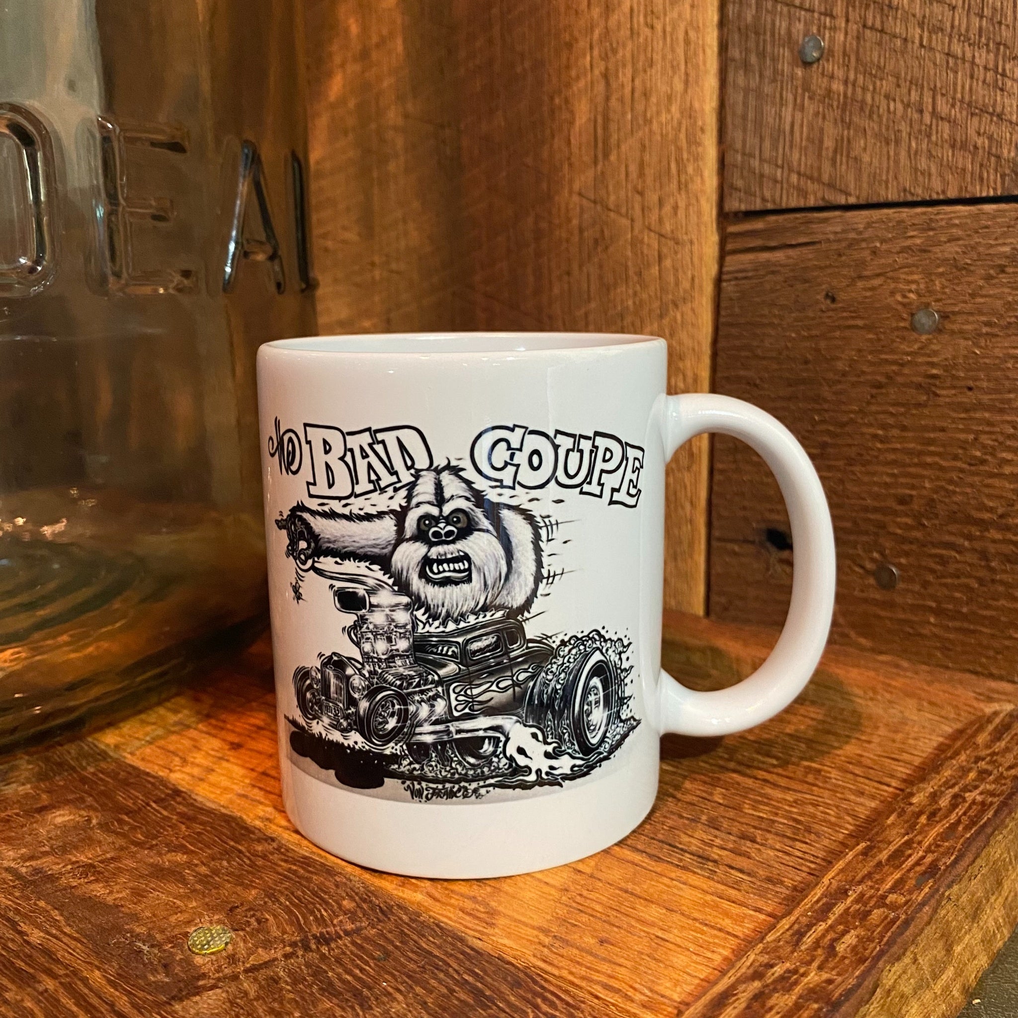 A white ceramic coffee mug with a black hand-drawn graphic of a cartoon creature driving a hot rod, with the text 'the Bad Coupe' above it. The mug is placed on a rustic wooden surface with a wooden wall background.